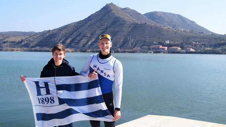 Die Hanseaten Felix Heinemann (links) und Johannes Rentz trainieren im spanischen Mequinenza für die anstehende U23-Saison. (Foto: Ruderclub Hansa von 1898 e.V. Dortmund) Die Hanseaten Felix Heinemann und Johannes Rentz halten eine Hansa.Flagge in die Kamera